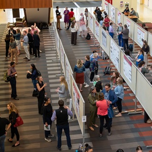 Top-down view of a busy academic poster session, with dozens of people clustered, talking around academic posters.